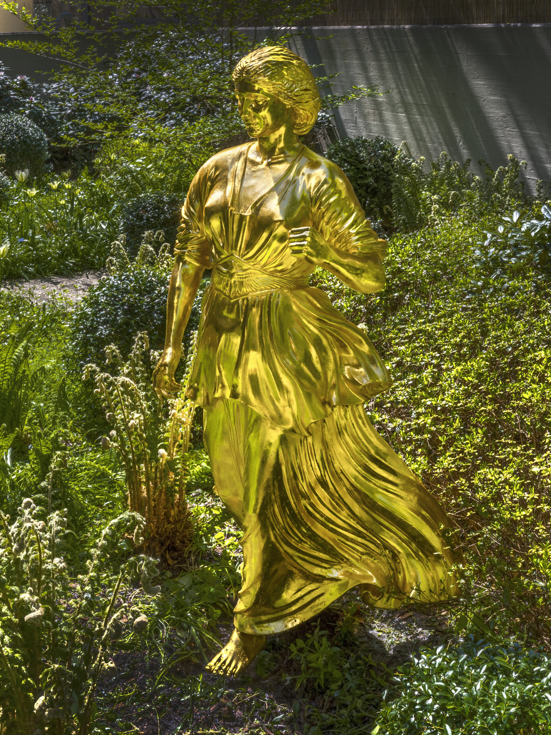 Goldene, weibliche Skulptur aus Bronze mit wallendem Kleid vor grüner Hecke